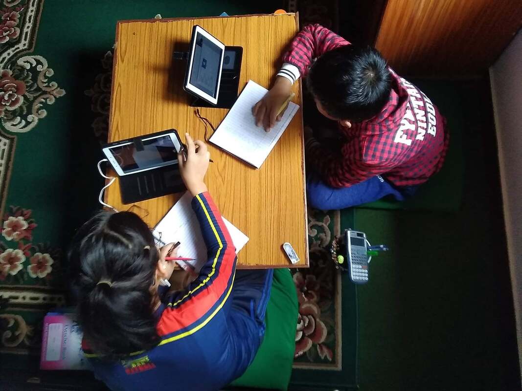 A girl and a boy in Nepal sit on the floor at a low table. They are using tablets to attend virtual school classes. They have notebooks, textbooks and a calculator around them.